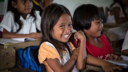 En liten flicka sitter i en skolbänk och ler mot kameran